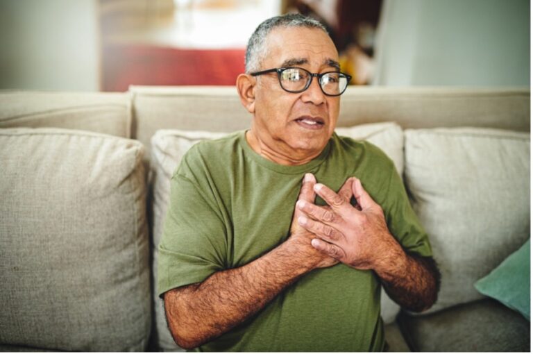 A senior man sits on the sofa holding his chest in pain caused by consuming a high level of fructose